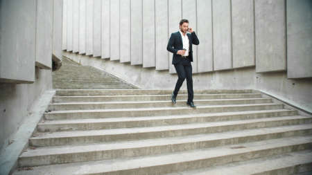 Handsome business man running down stairs in city. Busy male employee talking on smartphone outdoors. Confident businessman walking on city street with take away coffee. Man drinking coffee to goの写真素材