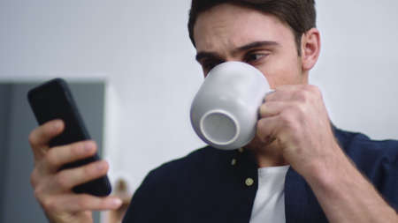 Portrait of young man typing mobile phone at home kitchen. Focused guy chatting on smartphone indoors. Closeup male person drinking coffee at modern apartment in slow motionの写真素材
