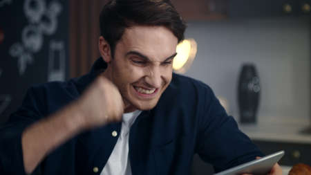 Portrait of excited man getting good news on tablet computer in home office. Cheerful businessman...