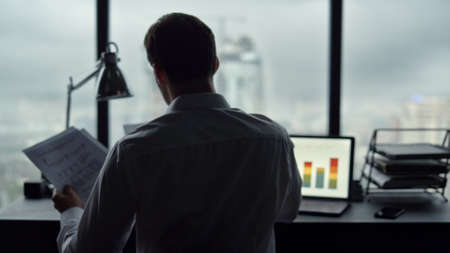 Back view unrecognizable businessman looking at bad statistics on papers. Worried professional touching head with hands at workplace. Sad business man sitting at table in office with laptopの写真素材