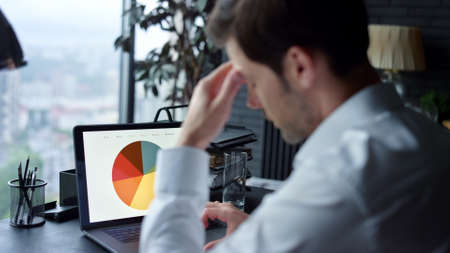 Sad businessman working on laptop with graphics in office. Upset male manager looking at financial...