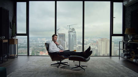 Handsome businessman relaxing in office chair with city view. Exhausted male employee adjusting tie in office interior. Tired business man sitting in office chair after work. Man sleeping in chairの写真素材