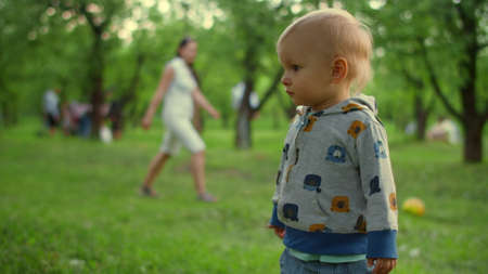 Close up focused toddler standing outside. Little boy running to family in forest. Happy mother and son playing with ball on background in park in slow motion.の写真素材