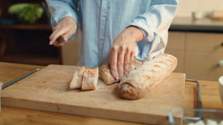 Closeup woman hands slicing bread with knife on wooden board. Attractive brunette female eating tasty bread in kitchen. Joyful girl enjoying smell of morning baguette in slow motion.の写真素材