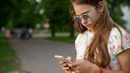 Close up cheerful girl playing games on mobile phone in park. Playful child using smartphone outdoors. Joyful teen girl texting in smartphone in slow motion.の写真素材