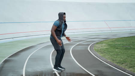 Active afro runner jumping on place at stadium. Male jogger warming before cardio workout on track....