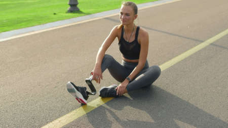 Smiling woman with artificial limb sitting on road. Cheerful girl relaxing after training in park....
