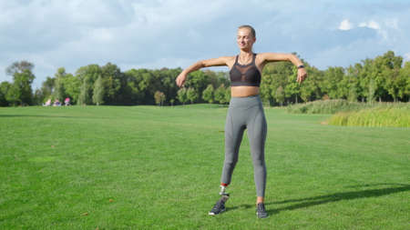 Sportswoman with artificial limb warming before workout in summer park. Fitness woman warming arms...