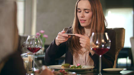 Businesswoman taking food photo on smartphone at restaurant. Smiling woman photographing dish on mobile phone in cafe. Business woman making photo of food at business lunchの写真素材
