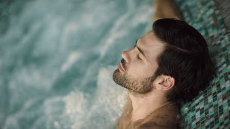 Close up of sexy man enjoying hot tub spa in slow motion. Portrait of handsome guy relaxing in pool at wellness hotel. Attractive man having hydro massage in hot tub indoor.の写真素材