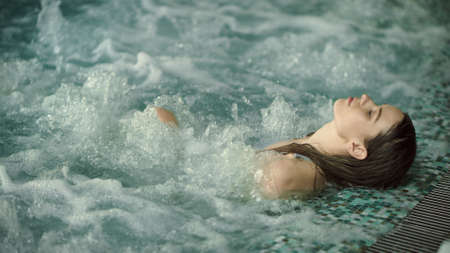Young woman relaxing in pool at luxury spa. Beautiful girl bathing at hot tub spa in modern hotel. Wet girl having hydrotherapy in luxury pool indoor.の写真素材