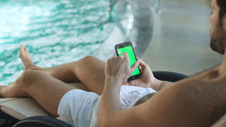 Closeup man hands holding mobile phone with green screen by pool at hotel. Close up of young man...