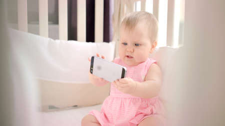 Little baby girl with phone in crib. Infant watching cartoonの写真素材