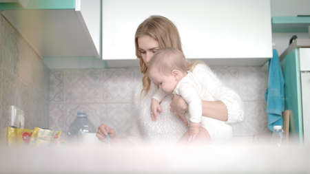 Woman with baby preparing food on kitchen. Mother with newborn child preparing breakfast at kitchen....