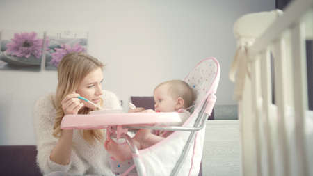 Beautiful mom try feeding baby with porridge. Blondy woman feed kid in baby chair at home. Family...