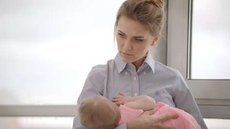Tired mother holding baby on hands near window. Stylish mom in shirt carrying baby. Upset businesswoman in business dress holding child on hands. Close up of tired woman holding kidの写真素材