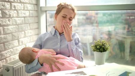 Tired woman yawn with sleeping child on hands. Tired mother yawning with baby and checking smartphone. Sleepy mom with kid at workplace. Yawning woman with baby at office workplace near windowの写真素材