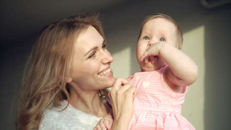 Happy mother holding baby girl on hands. Happy woman with baby on hands. Sweet motherhood. Portrait of mom with daugther at home. Cheerful mom embrance toddler girlの写真素材