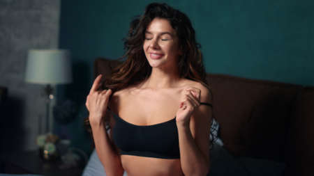 Portrait of sexy woman playing with bra straps in home bedroom interior. Closeup cheerful girl showing beckoning gesture with hand in bed. Sensual model girl flirting with camera in bed at hotel roomの写真素材