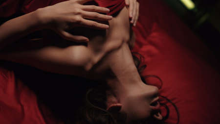 Closeup naked woman posing in bed under red silk sheet. Nude girl lying and touching body on satin linen bed. Sexy model girl caresses and excites herself with passion in bedroom.の写真素材