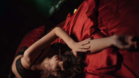 Top view seductive girl crossing hands on red silk sheets in bed. Closeup nude woman lying on satin bed. Charming model girl relaxing in sexy lingerie at dark bedroomの写真素材