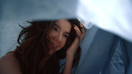 Portrait of pretty woman taking selfie photo by mobile phone under draped cloth blanket. Sexy girl using smartphone under bedsheet. Cheerful model girl posing under bed sheet.の写真素材