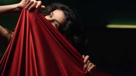 Portrait of sexy woman playing with red silk cloth. Closeup nude girl taking erotic poses with silk sheet indoors. Seductive girl flirting and posing for camera in dark bedroom.の写真素材