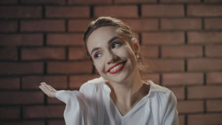 Young woman smiling and shrugging shoulders on brick wall copyspace. Happy woman laughing and posing on camera in studio. Cheerful girl portraitの写真素材