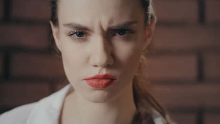 Beautiful woman with red lips frowning face posing in camera on brick wall background. Portrait scowling woman skeptical looking into camera in brick studio. Doubt, mistrust emotionsの写真素材