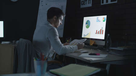 Overworked business man analyzing financial chart and diagram in night office. Business analyst looking economic data computer program in late office. Overtime work conceptの写真素材
