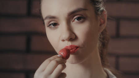 Sexy woman eating ripe strawberries on camera. Portrait sensual girl eating red strawberry and posing front camera on brick wall backgroundの写真素材