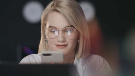 Happy business woman typing message on mobile phone in night office. Young business woman chatting in social networks at night work. Closeup girl working overtime at workplaceの写真素材