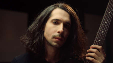 Portrait of young guitarist looking to camera in dark concert hall. Handsome composer holding...