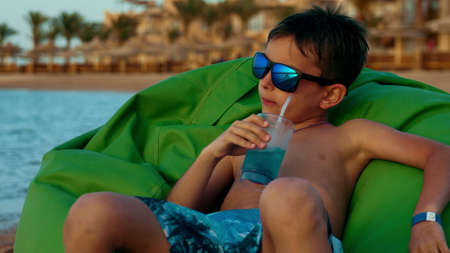 Teenage boy having fun in green soft chair at coastline in summer holiday. Young man drinking juice at sunny seashore. Attractive male person sitting at sunset seaside.の写真素材