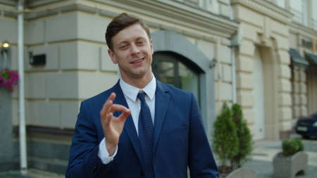 Portrait of confident business man looking at camera outdoors. Joyful man showing okay sign outside. Happy businessman standing at street. Closeup of cheerful professional posing at city streetの写真素材