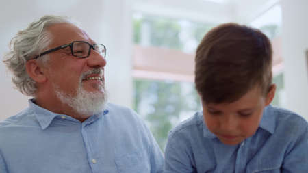 Smiling grandfather looking at grandson. Joyful boy sitting on grandparent laps. Portrait of adorable boy and senior man hugging each other at home. Family sitting on couch in living roomの写真素材