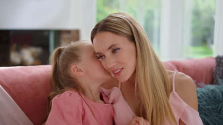 Smiling mother talking with daughter at home. Portrait of small girl whispering secret on woman ear....