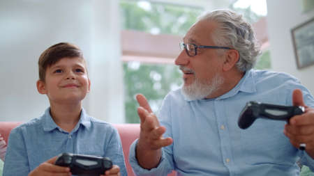 Focused senior man and boy playing video game with joysticks at home. Happy grandfather celebrating victory in game. Smiling grandparent and grandchild shaking hands after computer game overの写真素材