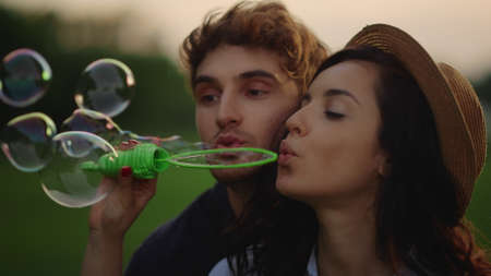 Portrait of playful couple blowing soap bubbles on meadow. Closeup happy man and woman having fun at...