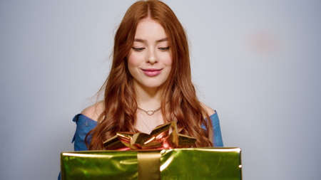 Closeup excited woman holding gift on gray background. Portrait of smiling female person shaking present box indoors. Surprised girl trying to guess box content in studio.の写真素材