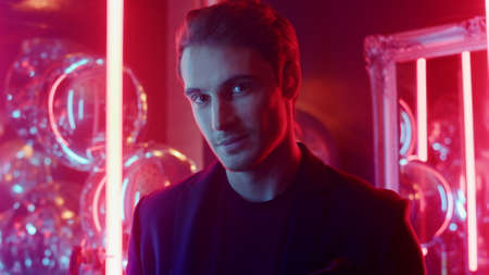 Closeup shy guy looking at camera in nightclub. Portrait of handsome male person posing on neon lights background. Young man smiling at lens at club party.の写真素材