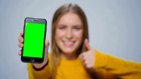 Closeup smiling woman showing smartphone green screen in studio. Portrait of happy girl holding mobile phone chroma key screen. Successful girl thumbs up cellphone mockup screen on grey background.の写真素材