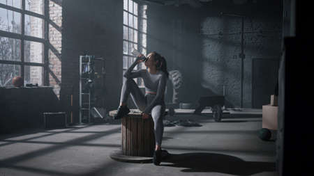 Tired girl drinking water in sports bottle at fitness training in dark gym. Female athlete refresh...
