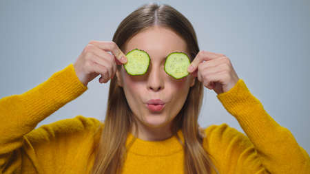 Portrait smiling woman having fun with two cucumber slices in studio. Closeup joyful girl making funny faces on grey background. Emotional model playing vegetable chunk. Healthy food diet skin careの写真素材