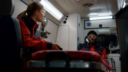 Medical team talking about work in emergency vehicle. Mixed race ambulance doctors driving on call. African american assistant talking with colleague. Female paramedic reading message on smartphoneの写真素材