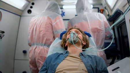Injured man lying on stretchers in emergency vehicle. Male victim breathing in oxygen mask. Patient feeling bad of coronavirus disease. Paramedics in protective suits working on backgroundの写真素材