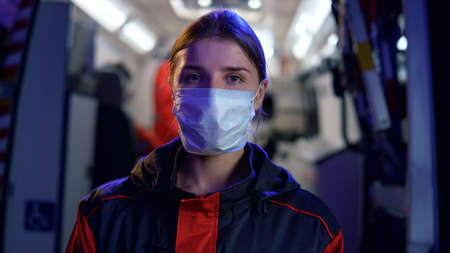 Professional paramedic in medical mask posing at camera. Portrait of woman wearing protective mask during coronavirus pandemic. Emergency care assistant standing at ambulance car on street at nightの写真素材