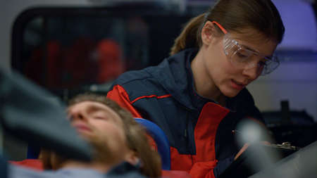 Male patient lying on stretchers in emergency car. Female paramedic in uniform writing disease diagnosis of victim on clipboard. Smiling ambulance doctor asking man about health conditionの写真素材