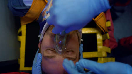 Top view paramedic hands in gloves putting oxygen mask on patient face. Unrecognizable ambulance doctors giving first aid help to injured man. Closeup victim in neck collar lying on stretchersの写真素材
