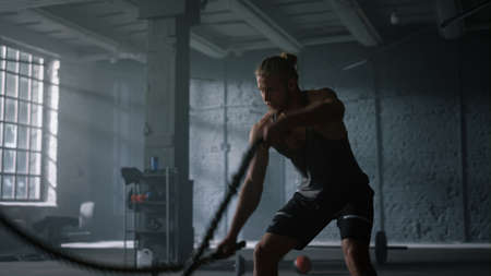 Muscular man training with battle ropes in modern gym. Excited male athlete doing intensity fitness...